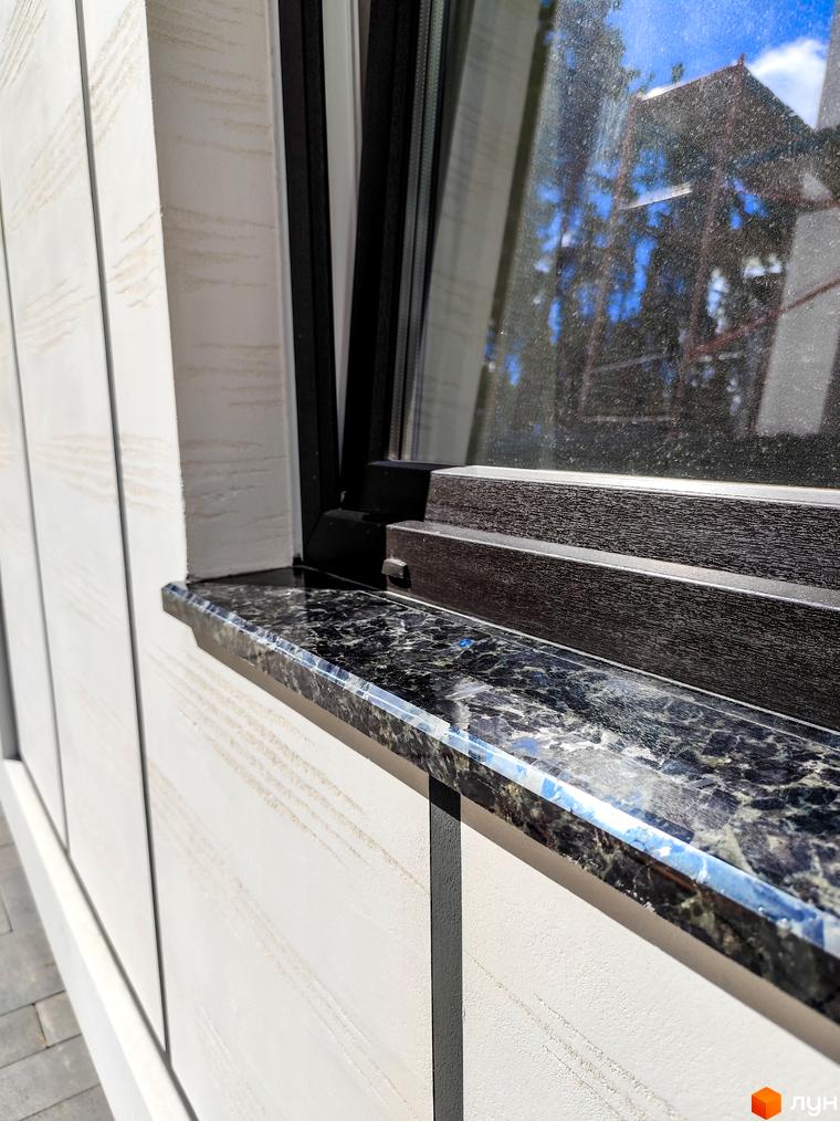 Вікно з темною рамою та підвіконням з чорного граніту на фоні білої фасадної панелі.