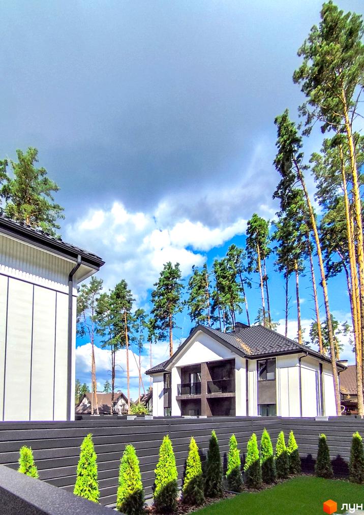 Сучасні дуплекси Toscana Home з чорним дахом та білими фасадами, оточені високими соснами та акуратним парканом з туями.