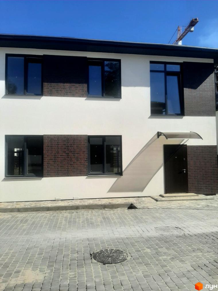 Котеджне містечко КМ Лісове-3: двоповерховий будинок з білою та цегляною обробкою, великими вікнами та темними дверима. Поруч вимощена плиткою територія та каналізаційний люк.