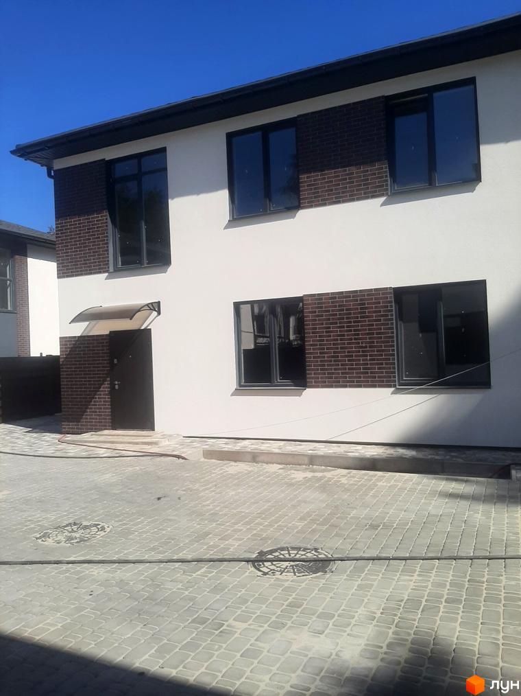 Зовнішній вигляд двоповерхового будинку з цегляними вставками та великими вікнами. Прибудинкова територія вимощена бруківкою з люками.