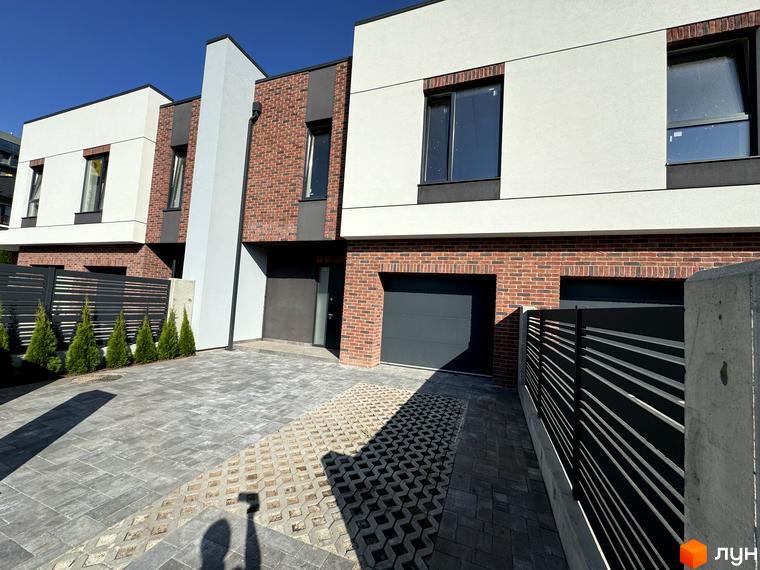 Таунхауси Loft House з цегляним фасадом та білими елементами, перед будинками розташовані гаражні ворота та вимощена сірими плитками прибудинкова територія з зеленими насадженнями.