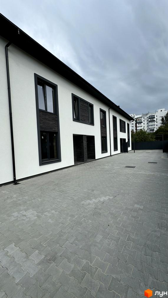 Таунхауси А56 з білою фасадною штукатуркою, чорними вікнами та дерев'яними елементами. Прибудинкова територія вимощена сірою плиткою.