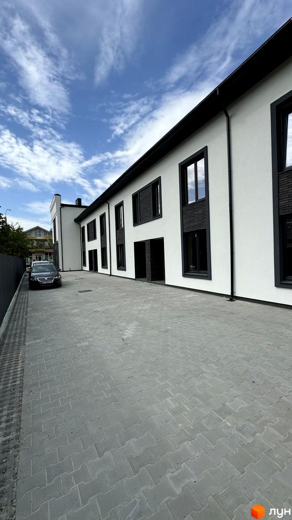 Таунхауси А56: біла двоповерхова будівля з темними вікнами та входами. Прибудинкова територія з бруківки та припаркований автомобіль.