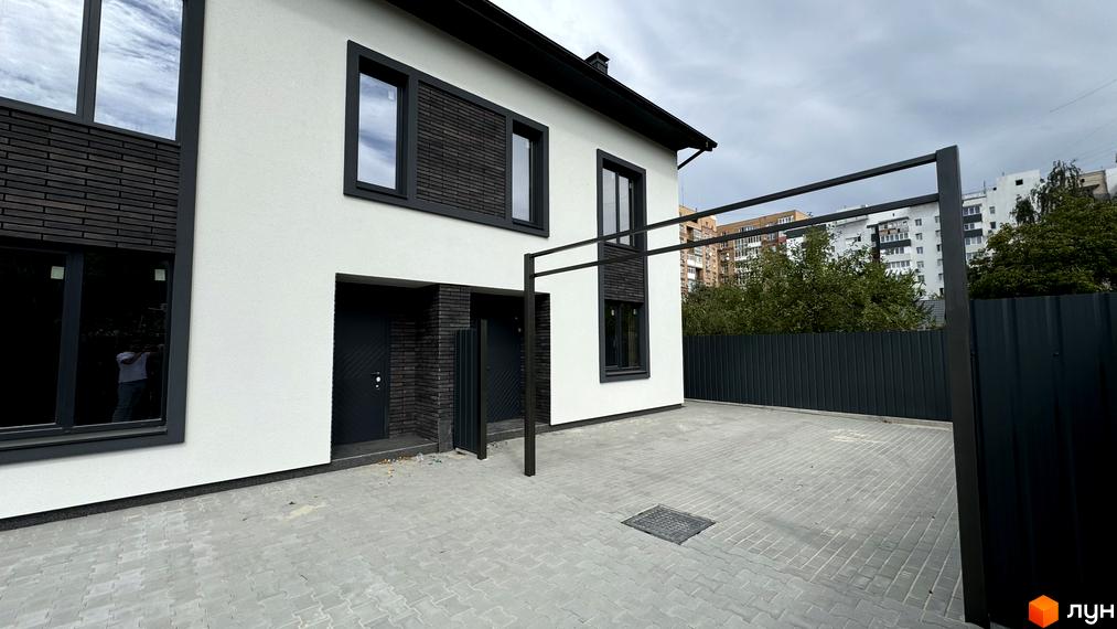 Таунхаус А56 з білою штукатуркою, темними вікнами та цегляними вставками, на тлі паркану та сусідніх будинків. Прибудинкова територія вимощена плиткою.