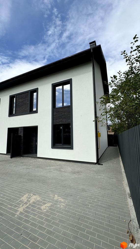 Таунхаус А56 з білою стіною, чорними вікнами та дахом, розташований на вимощеній сірою плиткою території з парканом.