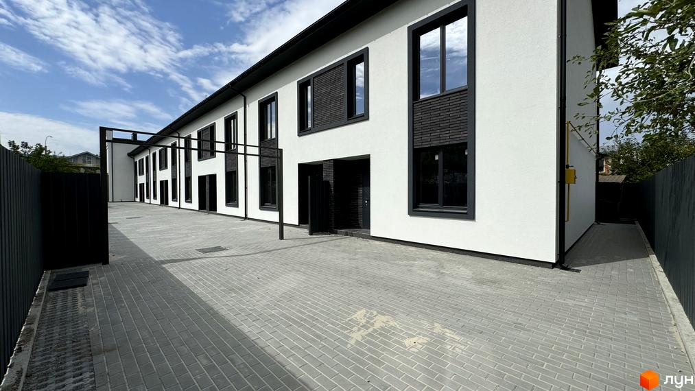 Зовнішній вигляд таунхаусів А56 з білою штукатуркою та темними вікнами. Прибудинкова територія вимощена плиткою.