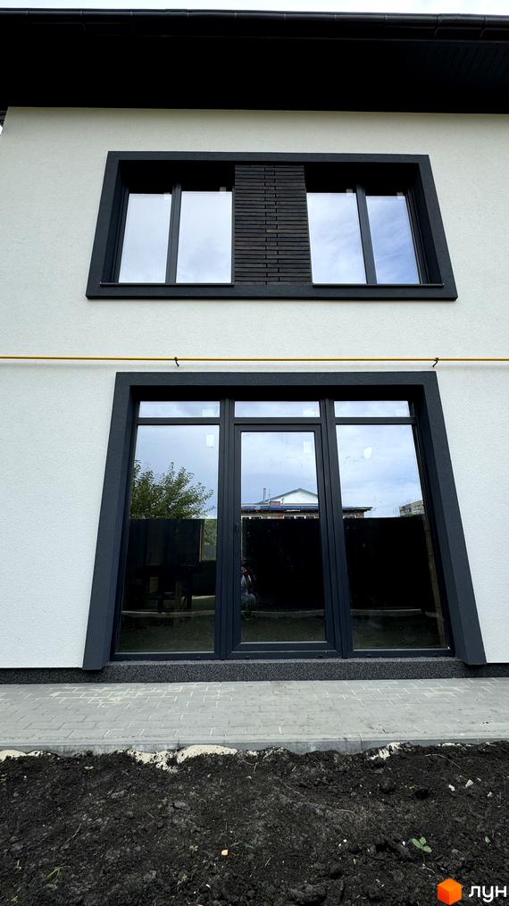 Таунхауси А56: фасад будинку з великими темними вікнами та сірою штукатуркою, вимощення з плитки та ділянка з землею.