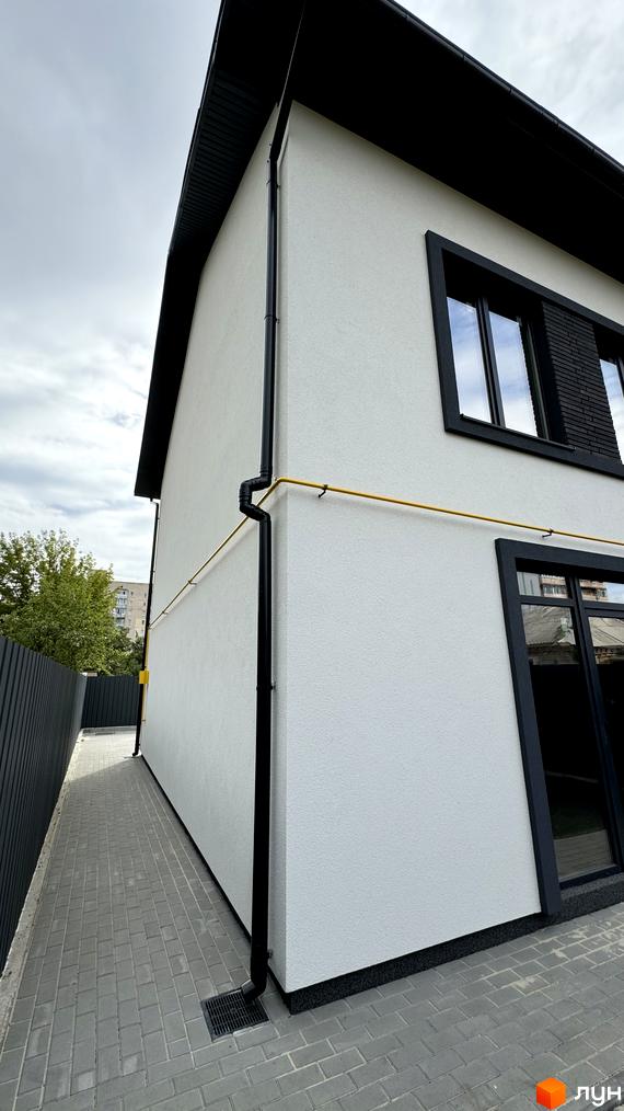 Зовнішній вигляд таунхауса А56 з білою штукатуркою та чорними вікнами. Прибудинкова територія з бруківкою та парканом.