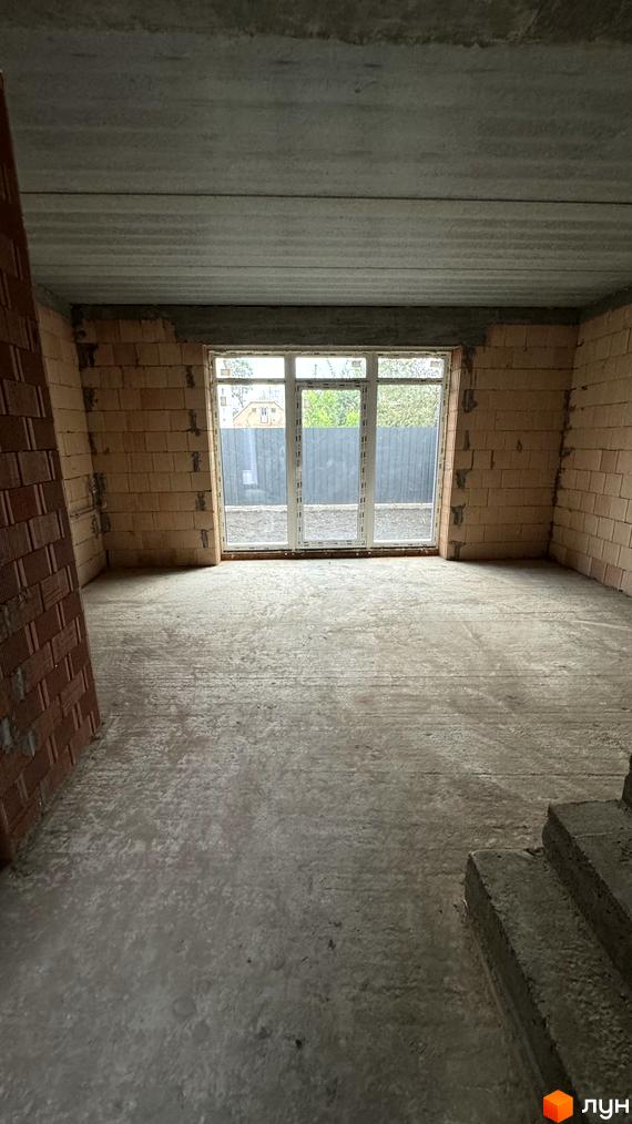 Таунхауси А56: вид зсередини на незавершений ремонт з цегляними стінами та великим вікном. Зовні видно паркан та дерева.