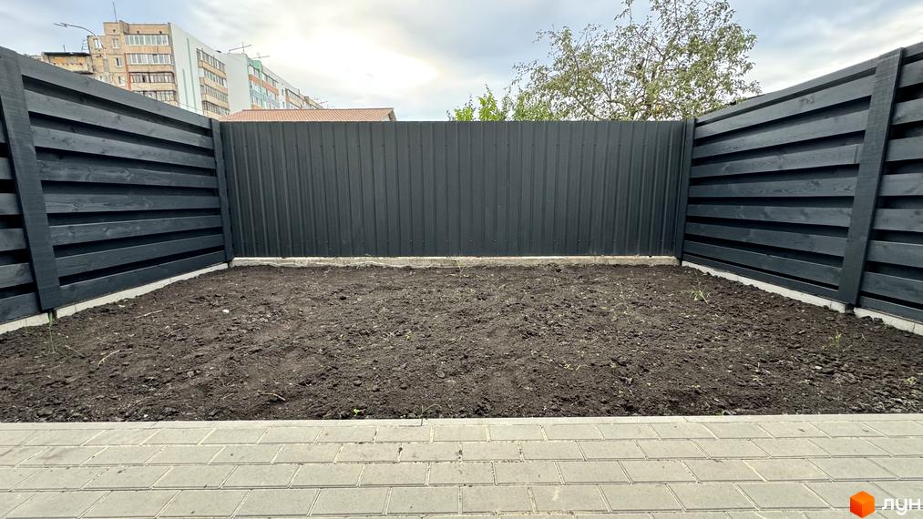 Таунхауси А56 з чорним дерев'яним парканом та ділянкою землі. На задньому плані видно багатоповерхові будинки та дерева.