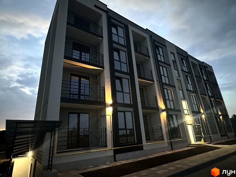 Зовнішній вигляд будинку ЖК Brickwood 2 з балконами та вікнами, освітлений вечірніми ліхтарями. Навколо будинку облаштована прибудинкова територія з доріжками.