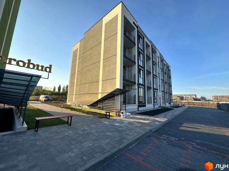 Зовнішній вигляд новобудови ЖК Brickwood 2 з сірими та білими фасадами та балконами. Поруч розташована прибудинкова територія з лавками та доріжками.