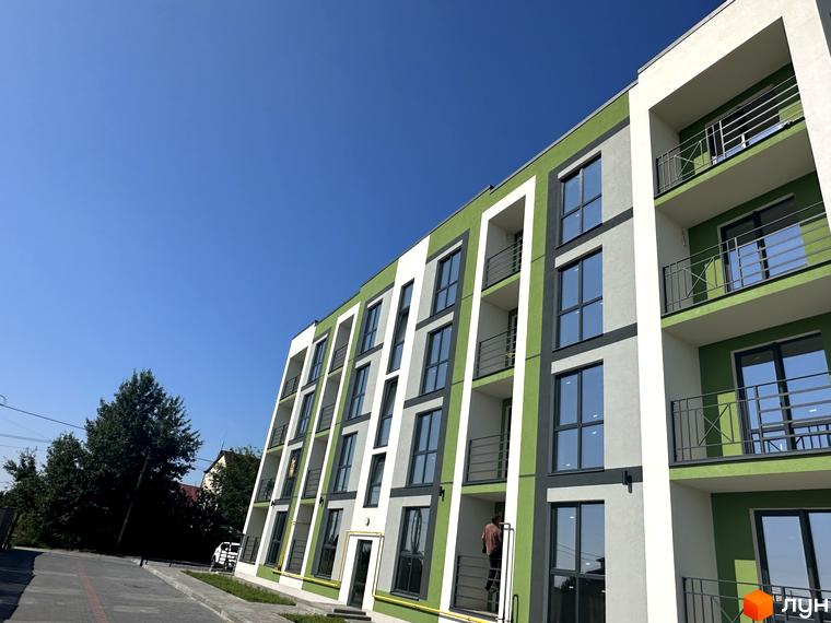 Зовнішній вигляд житлового комплексу Brickwood 2 з зеленими та білими фасадами, балконами та вікнами. Поруч розташована прибудинкова територія з деревами та дорогою.