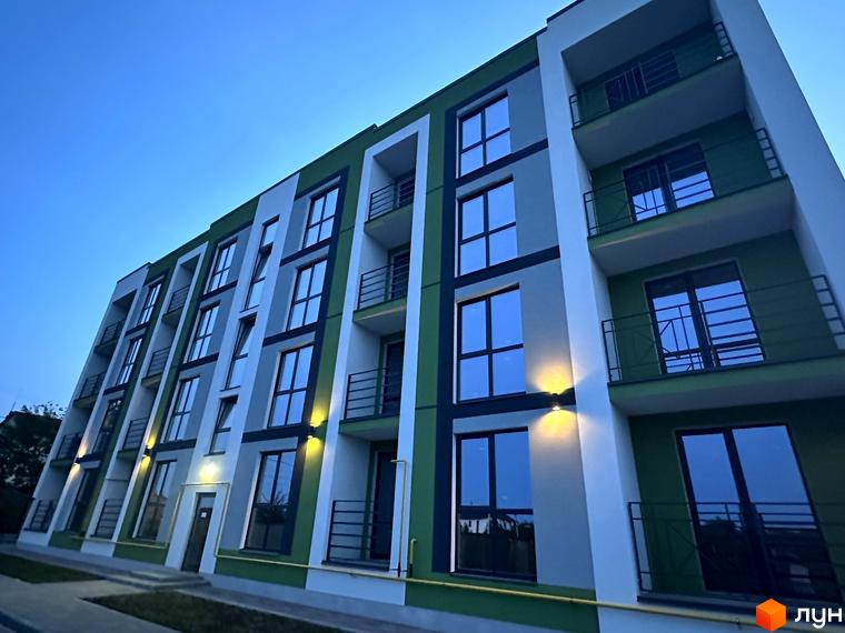 Фасад житлового комплексу Brickwood 2 з великими вікнами та балконами, освітлений вечірніми ліхтарями. Навколо будинку видно зелену зону та тротуар.