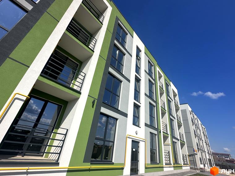 Зовнішній вигляд новобудови ЖК Brickwood 2 з фасадом у біло-зелених тонах та балконами. Навколо будинку видно елементи прибудинкової території та ясне блакитне небо.