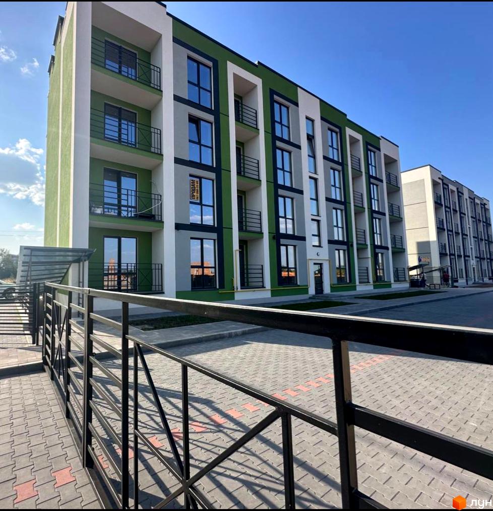 Зовнішній вигляд новобудови ЖК Brickwood 2 з зеленими та сірими фасадами, вікнами та балконами. Прибудинкова територія з бруківкою та огорожею.