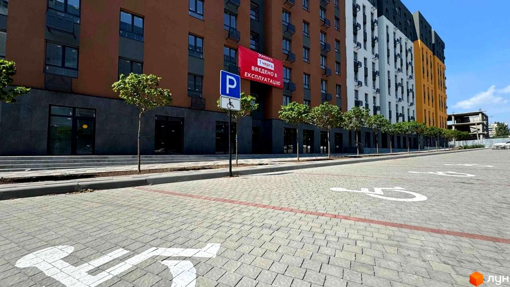 Житловий комплекс Osnova з оранжевим та сірим фасадами, парковкою з позначками для людей з інвалідністю та зеленими насадженнями.