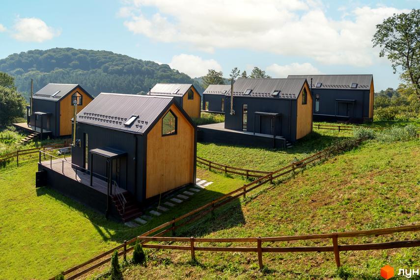 Котеджне містечко Схили: сучасні будинки з темними фасадами та дерев'яними елементами на тлі зелених пагорбів. Навколо будівель розташована доглянута прибудинкова територія з травою та дерев'яними парканами.