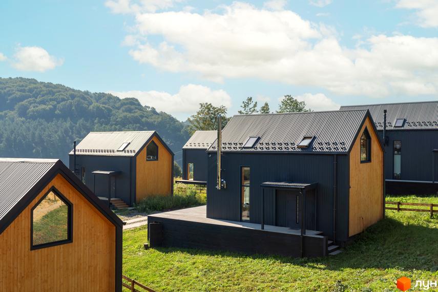 Котеджне містечко Схили: сучасні будинки з дерев'яними фасадами та темними дахами на тлі зелених схилів. Навколо будинків доглянута трава та дерева.