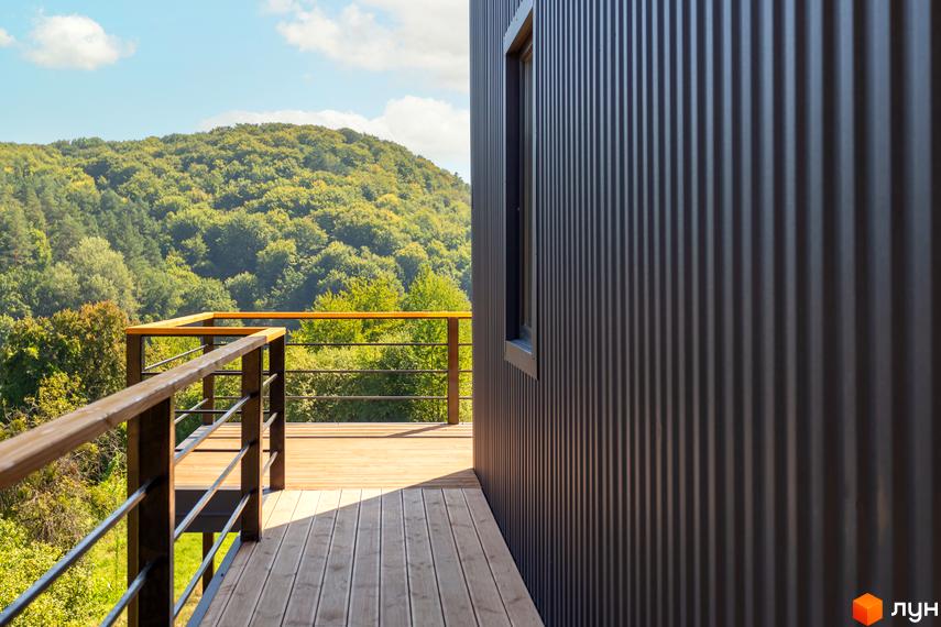 Зовнішній вигляд будинку з металевим фасадом та дерев'яною терасою. Навколо зелені пагорби та ліс.