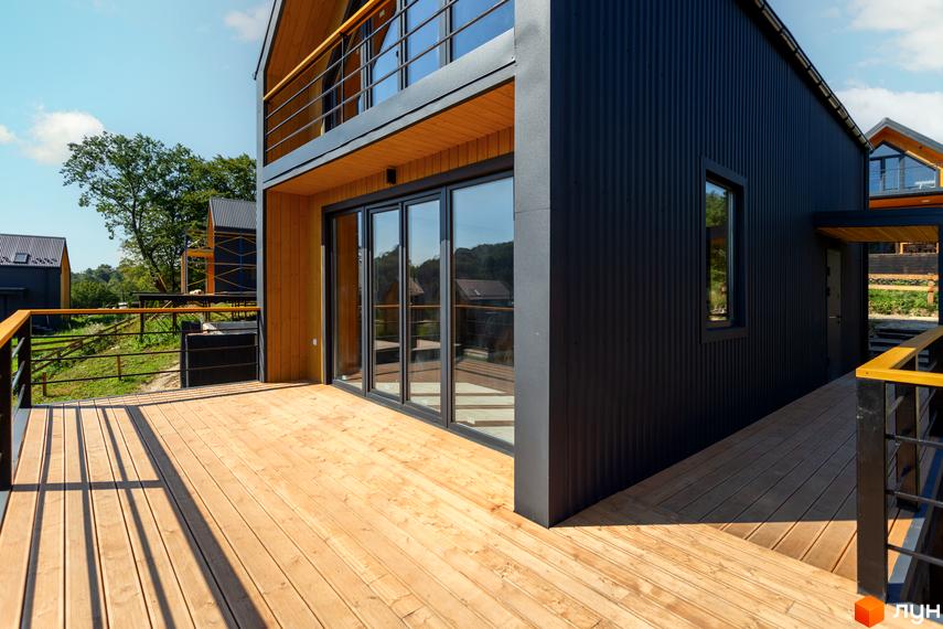 Котеджне містечко КМ Схили. Зовнішній вигляд будинку з дерев'яною терасою та чорним фасадом. Навколо зелена територія.