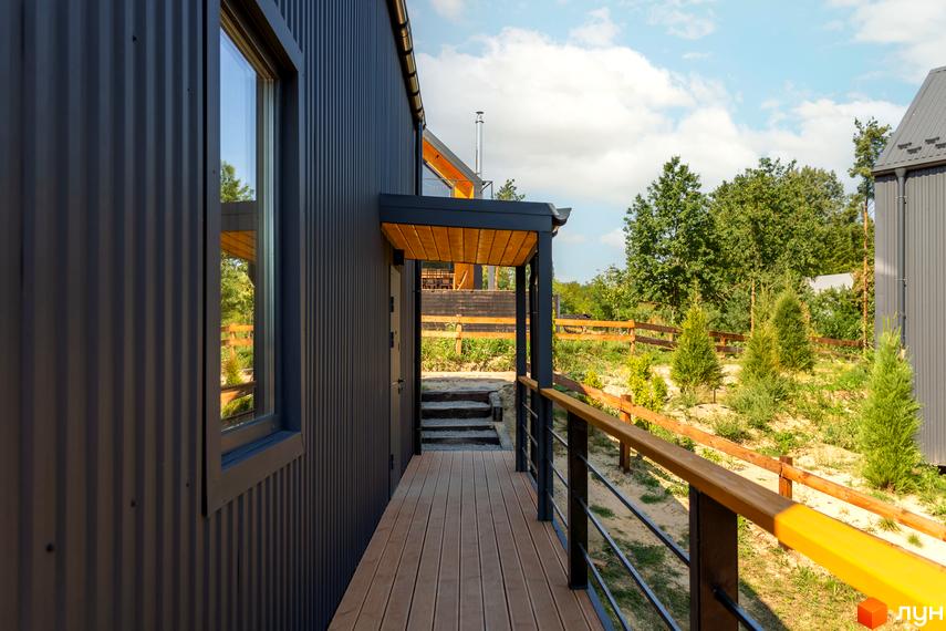 Зовнішній вигляд будинку з темним металевим фасадом та дерев'яною терасою. Прибудинкова територія з зеленими насадженнями та дерев'яним парканом.