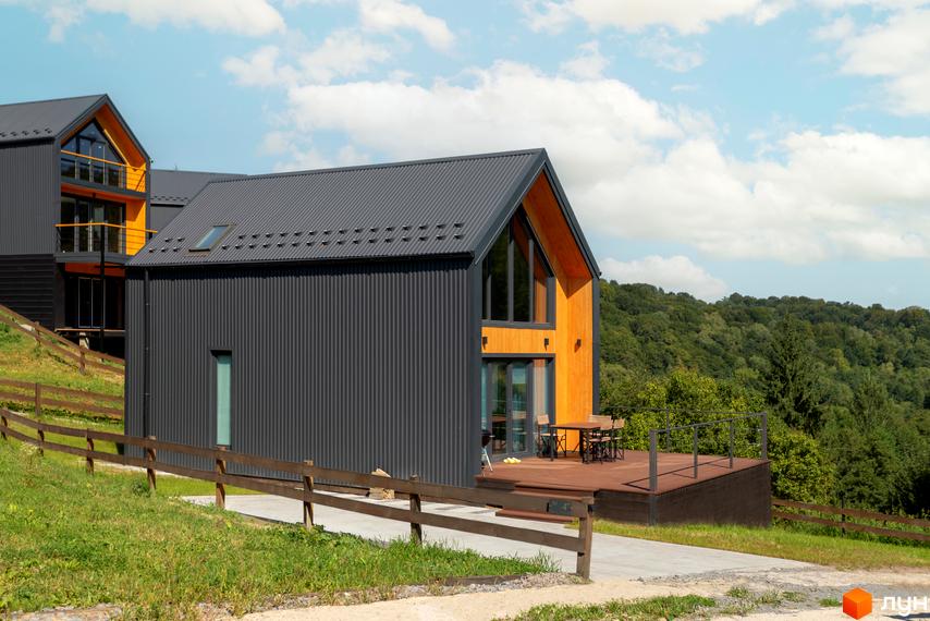 Котеджне містечко Схили: сучасні будинки з темним фасадом та дерев'яними елементами, розташовані на схилі пагорба з видом на ліс. Прибудинкова територія з дерев'яною терасою та огорожею.