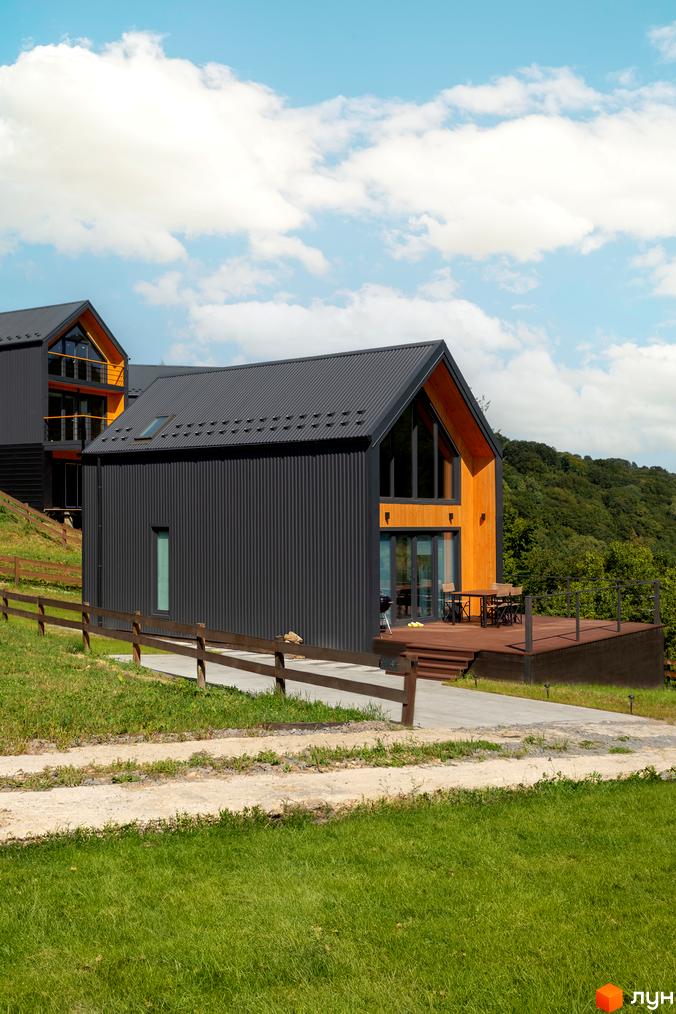 Котеджне містечко Схили: сучасні будинки з темним фасадом та дерев'яними елементами на тлі зелених схилів. Прибудинкова територія з доглянутим газоном та дерев'яною огорожею.