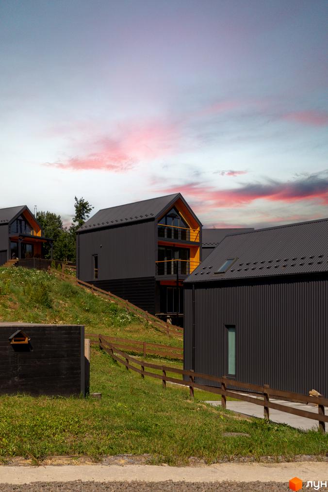 Котеджне містечко Схили: сучасні будинки з темним фасадом та жовтими акцентами на схилі пагорба, оточені зеленню та дерев'яним парканом.