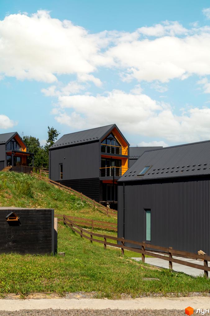 Сучасні будинки з темним фасадом та жовтими акцентами розташовані на схилі пагорба. Навколо зелена трава та дерев'яний паркан.
