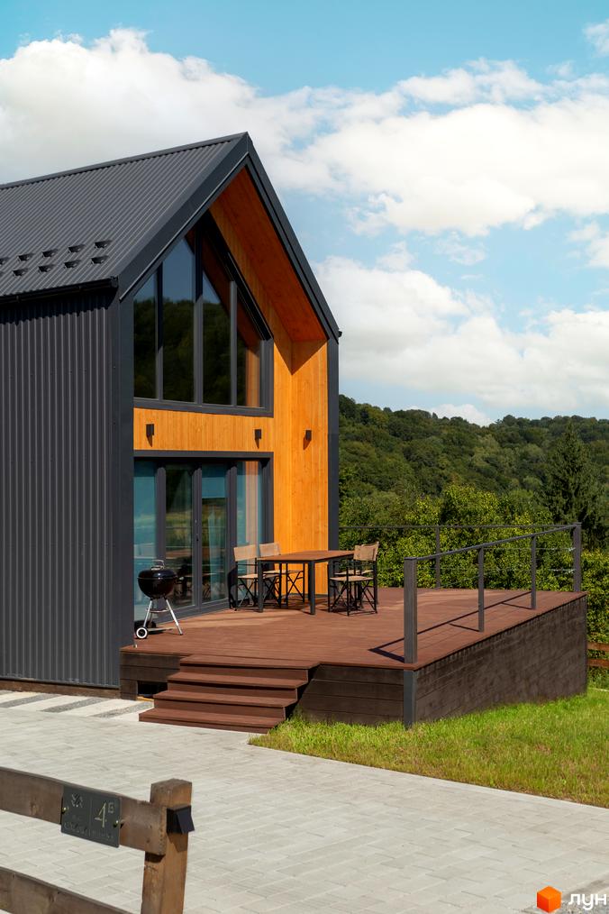 Котеджне містечко КМ Схили. Зовнішній вигляд будинку з дерев'яною обшивкою та великими вікнами, тераса з меблями та мангал.