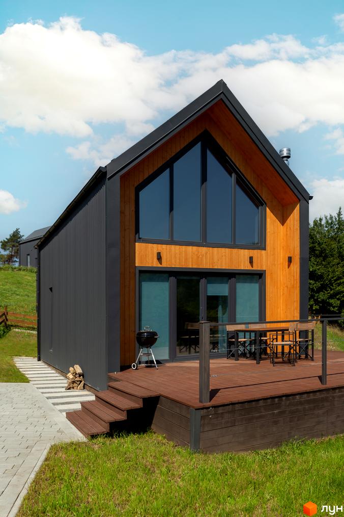 Котеджне містечко Схили: двоповерховий будинок з великими вікнами та дерев'яним фасадом, тераса з меблями та мангал. Навколо будинку зелена трава та доріжка з плитки.