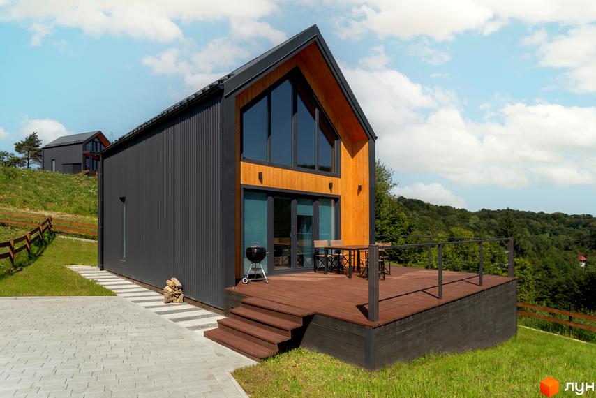 Котеджне містечко Схили: двоповерховий будинок з темним профнастилом та дерев'яними елементами, великими вікнами та терасою. Навколо будинку зелена ділянка з доріжкою з плитки.