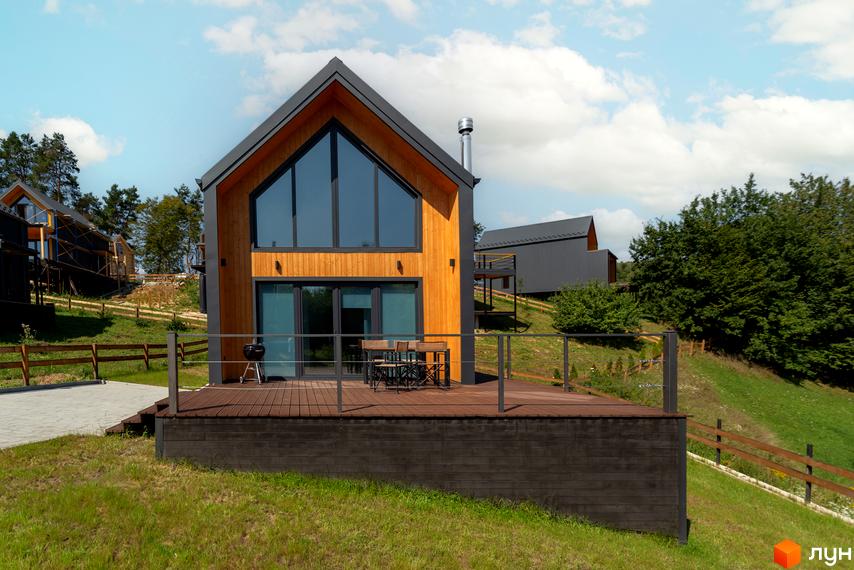 Котеджне містечко Схили: дерев'яний будинок з великими вікнами та терасою, оточений зеленню та іншими будинками на схилі.