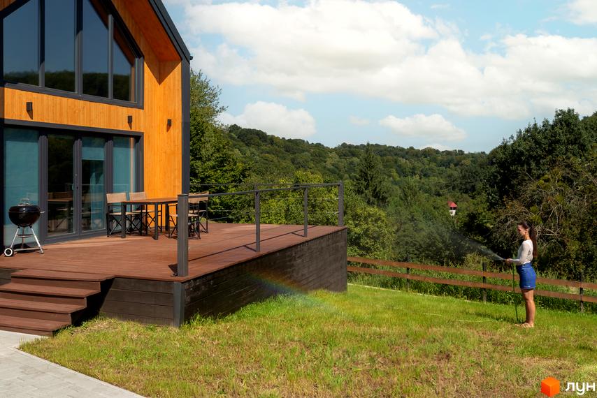 Котеджне містечко КМ Схили: двоповерховий будинок з дерев'яною обшивкою та великими вікнами, тераса з меблями та барбекю, зелений двір з огорожею та лісом на задньому плані.