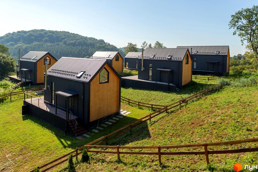 Котеджне містечко Схили: сучасні будинки з дерев'яним оздобленням та чорними фасадами на тлі зелених пагорбів. Територія комплексу огороджена дерев'яним парканом.