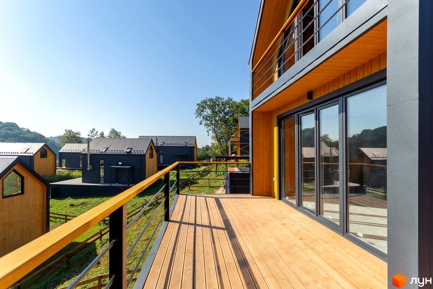 Котеджне містечко КМ Схили: дерев'яні та сірі будинки з великими вікнами та терасами на тлі зеленого ландшафту під блакитним небом.