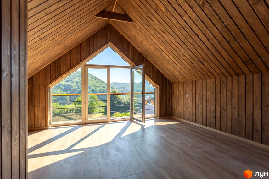 Інтер'єр горищного поверху з дерев'яними стінами та стелею, великим трикутним вікном та балконом з видом на зелені пагорби. Житловий комплекс КМ Схили.