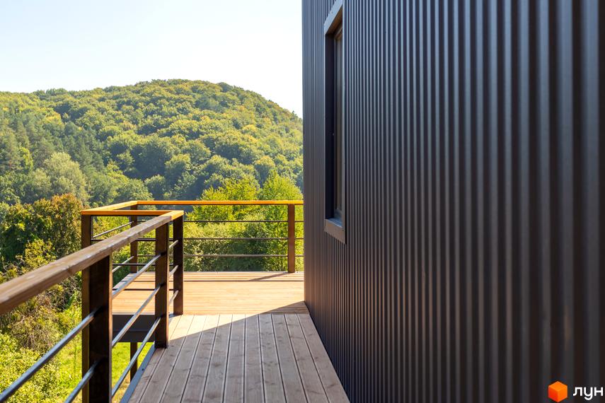 Котеджне містечко Схили: дерев'яна тераса з металевими перилами на тлі лісистого схилу та сучасної стіни будинку з вікном.