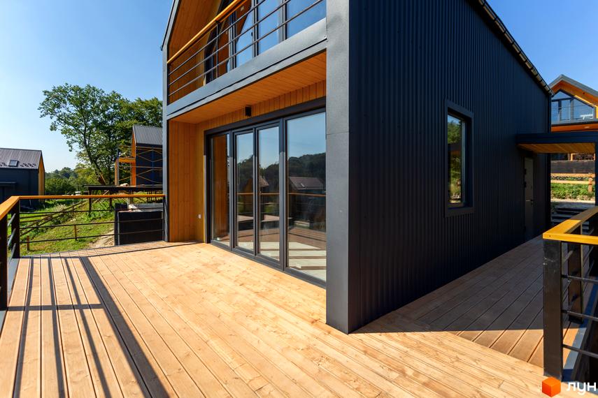 Котеджне містечко КМ Схили: двоповерховий будинок з дерев'яною терасою та чорним фасадом на тлі зеленого ландшафту.