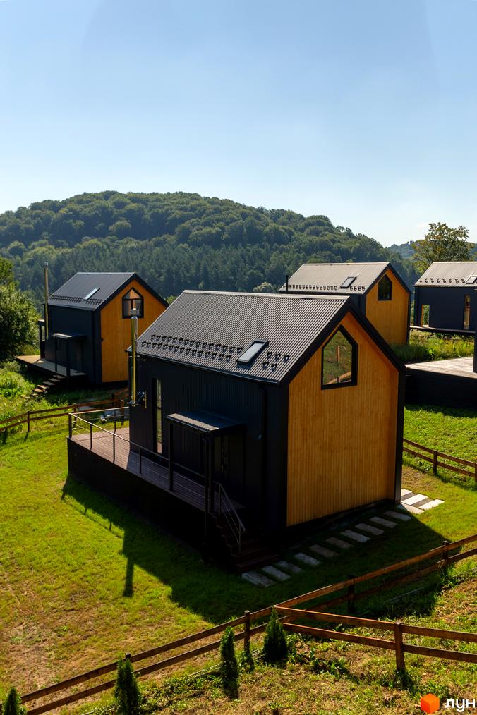 Котеджне містечко Схили: дерев'яні будинки з чорними дахами на тлі зелених пагорбів та блакитного неба. Територія комплексу вкрита травою та огороджена дерев'яним парканом.