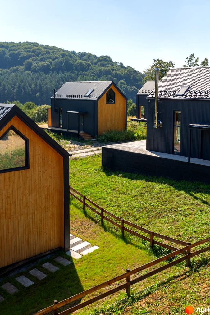 Котеджне містечко КМ Схили: сучасні будинки з дерев'яними фасадами та чорними дахами на тлі зелених пагорбів. Прибудинкова територія з газоном та дерев'яним парканом.