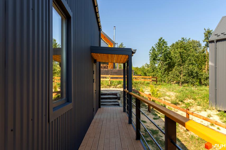 Зовнішній вигляд будинку з темно-сірого профнастилу з дерев'яною терасою та перилами. Прибудинкова територія з зеленими деревами та дерев'яним парканом.