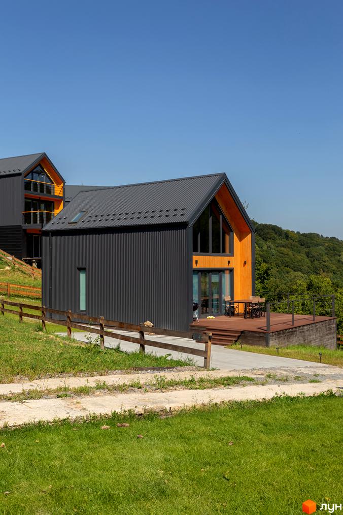 Котеджне містечко Схили: сірі будинки з дерев'яними елементами та терасою на тлі зелених пагорбів. Навколо будівель – доглянута територія з травою та дерев'яним парканом.