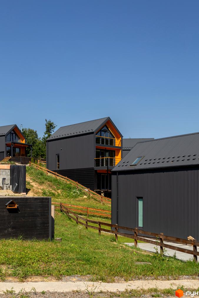 Котеджне містечко Схили: сірі будинки з темним дахом та жовтими елементами фасаду на фоні зеленого схилу з дерев'яним парканом.