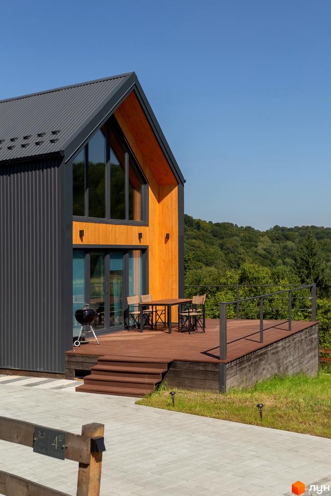 Котеджне містечко Схили: будинок з металевою та дерев'яною обшивкою, великими вікнами. Тераса зі столиком та стільцями на фоні лісу.