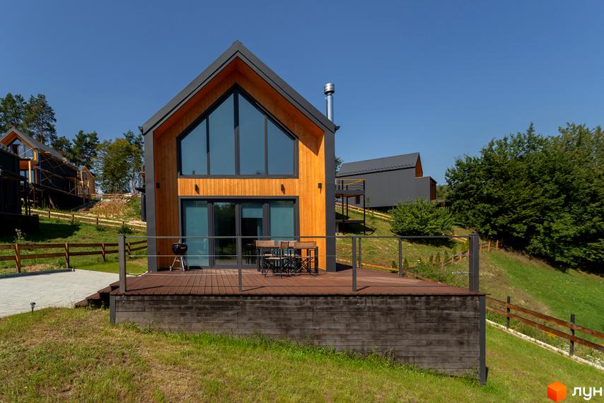 Котеджне містечко Схили: двоповерховий будинок з дерев'яним фасадом, великими вікнами та терасою. Навколо будинку зелена галявина та огорожа.