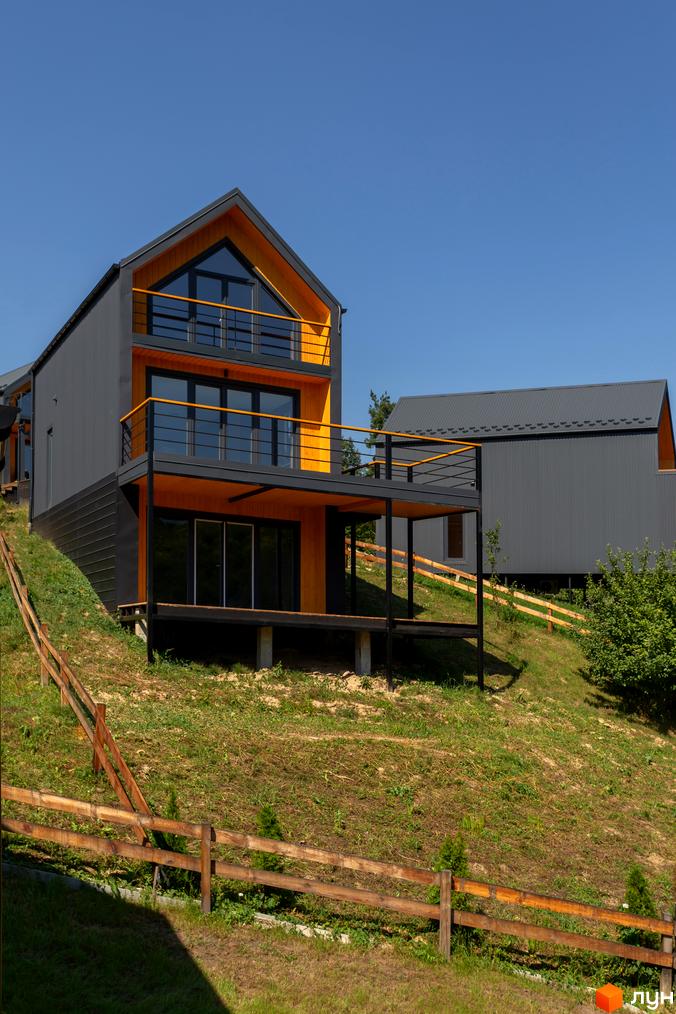 Котеджне містечко Схили: двоповерхові будинки з темними фасадами та жовтими акцентами на схилі гори. Навколо зелена трава та дерев'яний паркан.