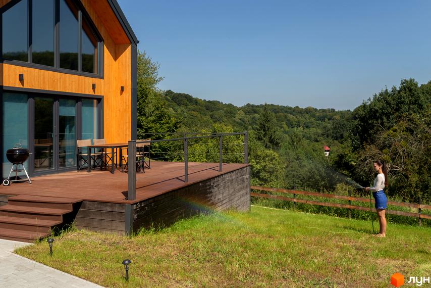 Котеджне містечко КМ Схили: дерев'яний будинок з великими вікнами та терасою, зелена галявина з поливом.