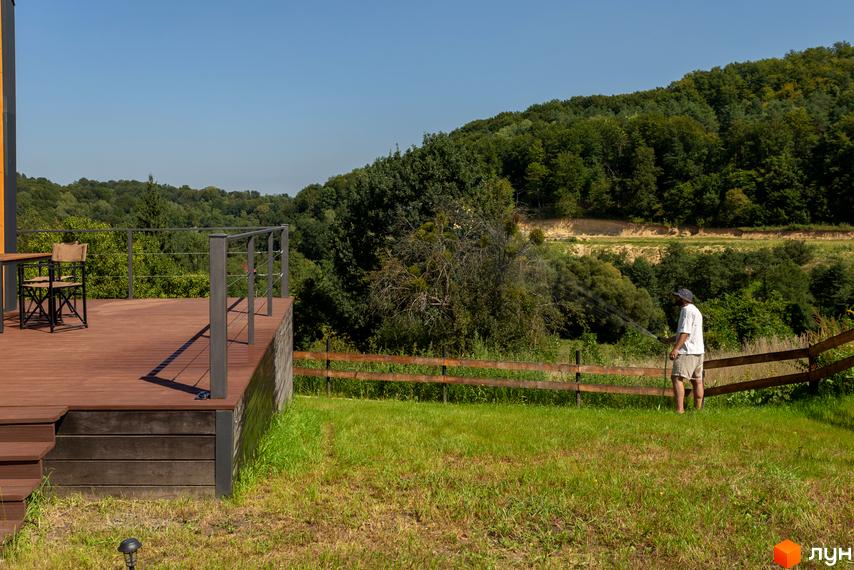 Котеджне містечко Схили: вид з тераси на зелений пагорб, де чоловік поливає газон.